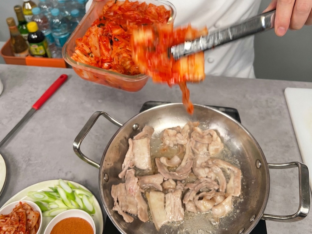 Duża porcja jaskrawoczerwonego kimchi przekładana ze szklanego naczynia na patelnię z podsmażonym boczkiem.