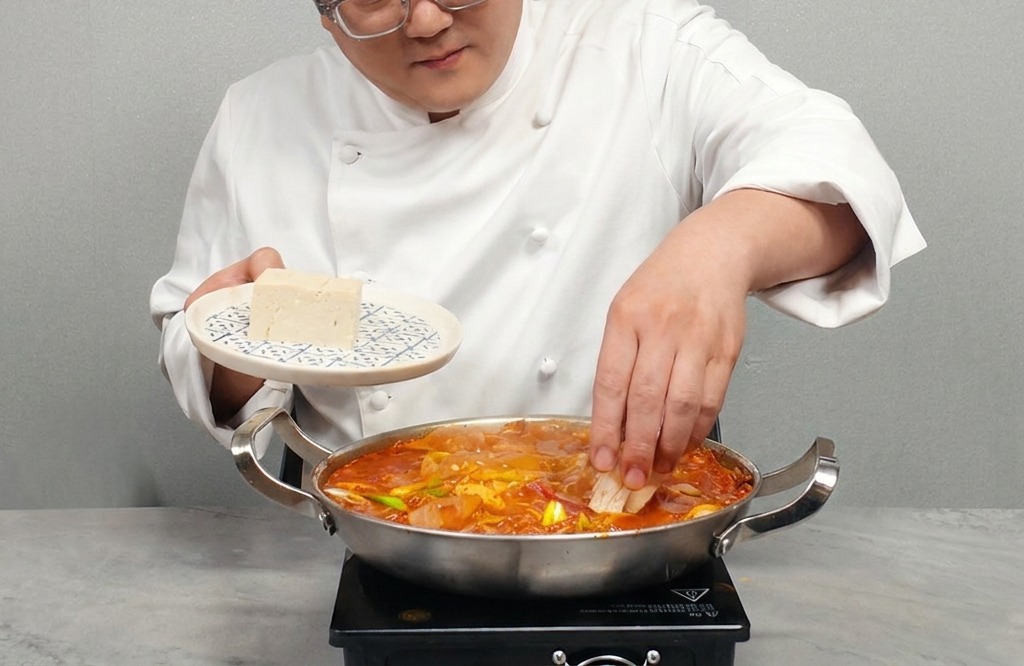 Osoba układająca plastry białego, twardego tofu w garnku z gorącym Kimchi Jjigae.