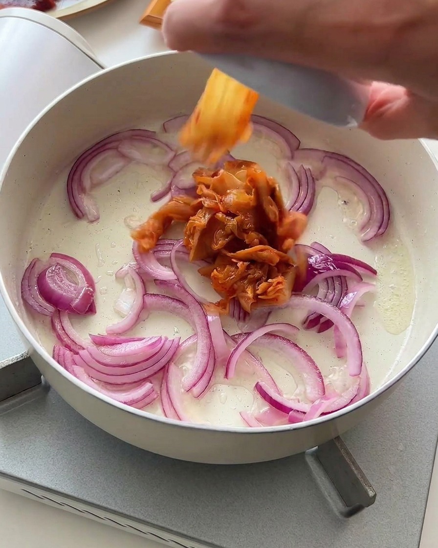 Posiekane kimchi dodawane do podsmażonej czerwonej cebuli na patelni.