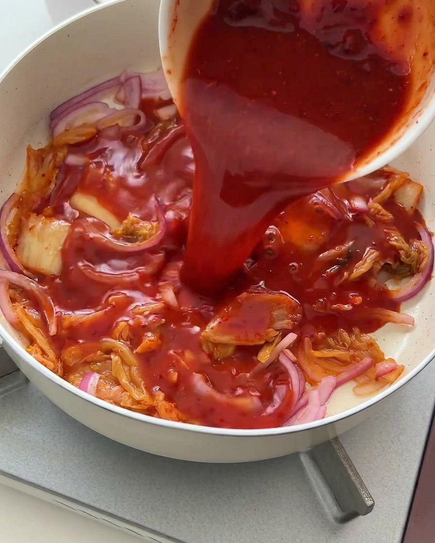 Intensywnie czerwony sos wlewany z miseczki na podsmażone kimchi i cebulę.