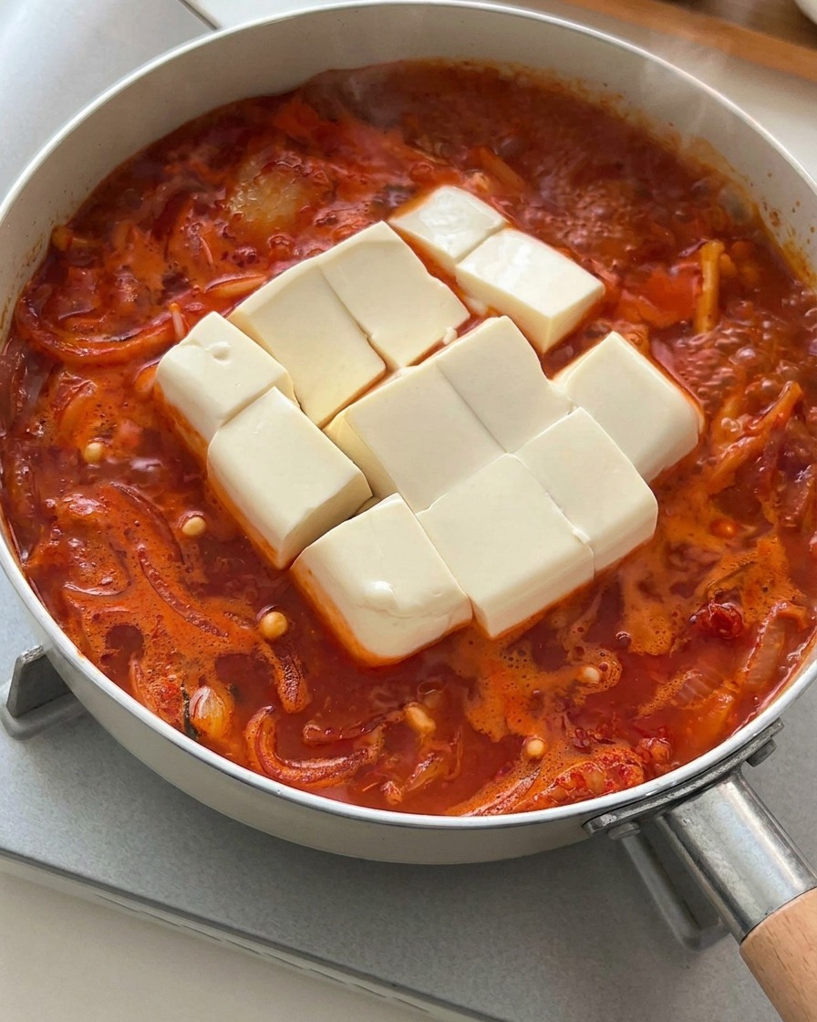 Blok miękkiego białego tofu, pokrojony w kostkę, wkładany do gotującej się zupy kimchi.