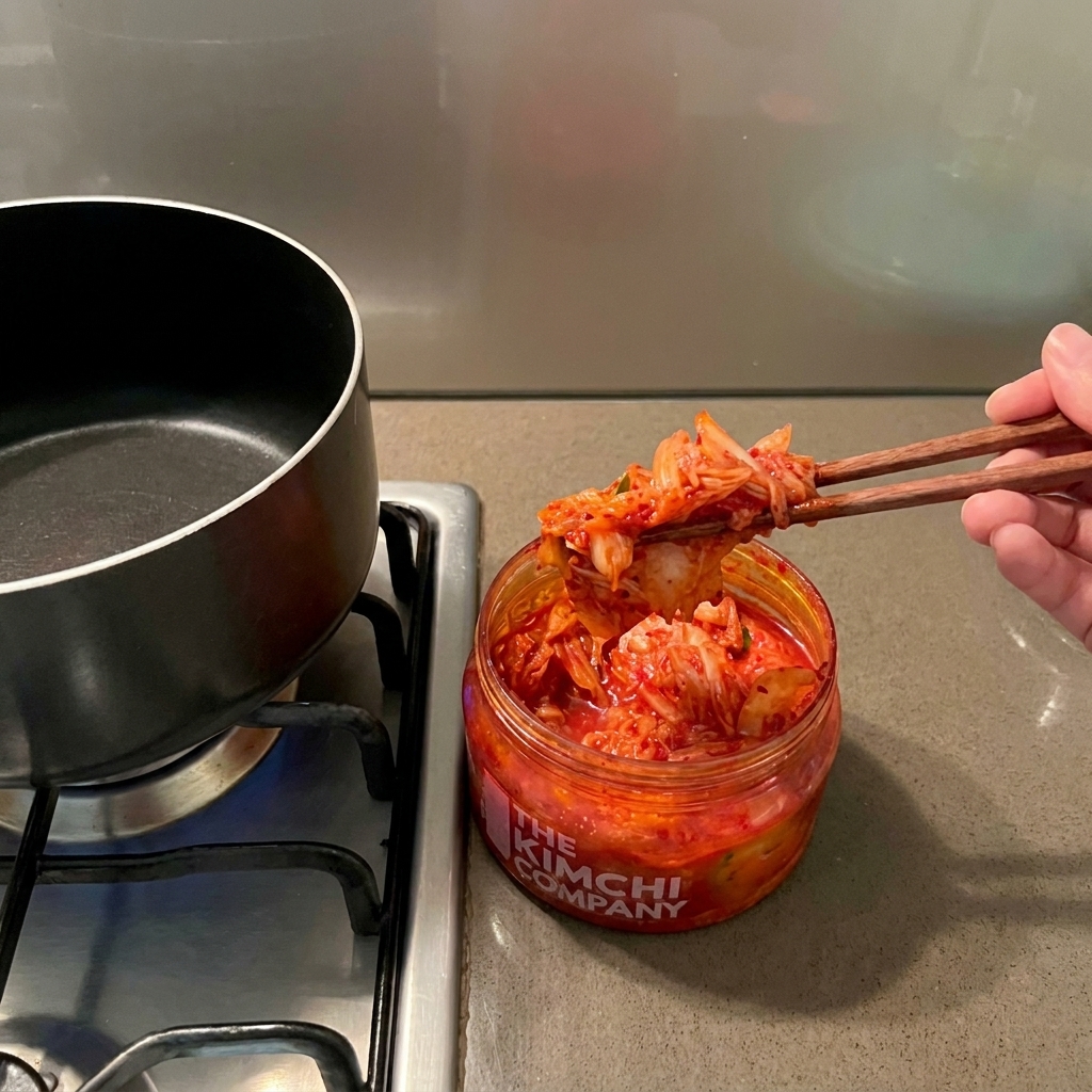 Ręka trzymająca drewniane pałeczki, wyjmująca czerwone kimchi ze słoika nad pustym czarnym garnkiem.