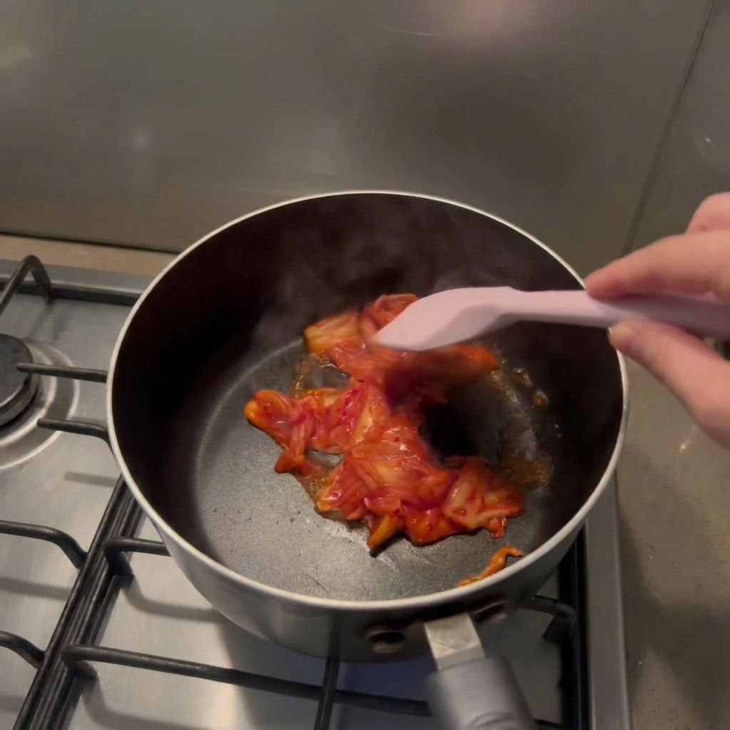 Ręka mieszająca różową silikonową szpatułką posiekane kimchi w czarnym garnku.
