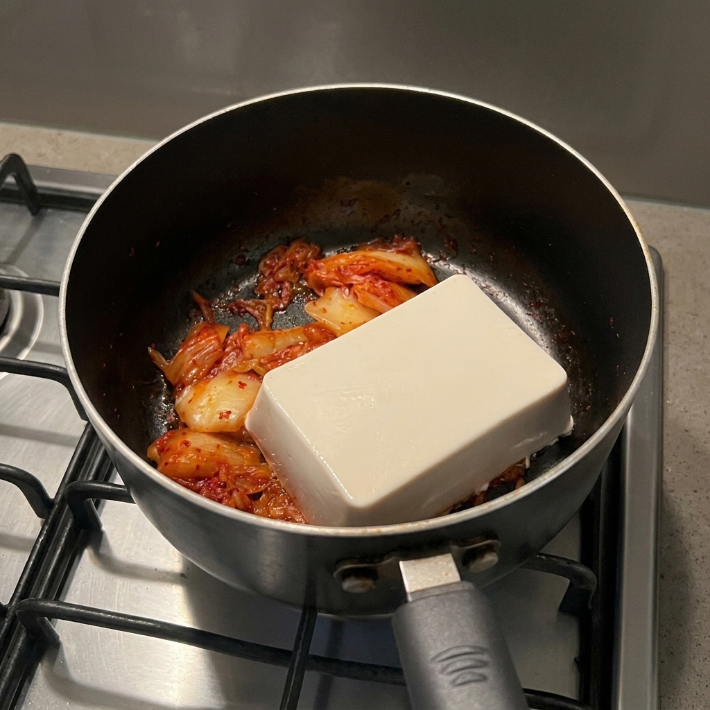 Prostokątny blok białego, miękkiego tofu leżący na podsmażonym kimchi w czarnym garnku.