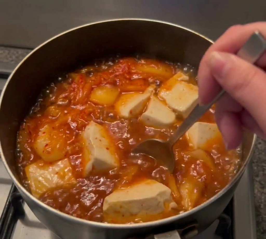 Metalowa łyżka mieszająca głęboko czerwoną pastę chili w bulgoczącym garnku z zupą, tofu i kimchi.