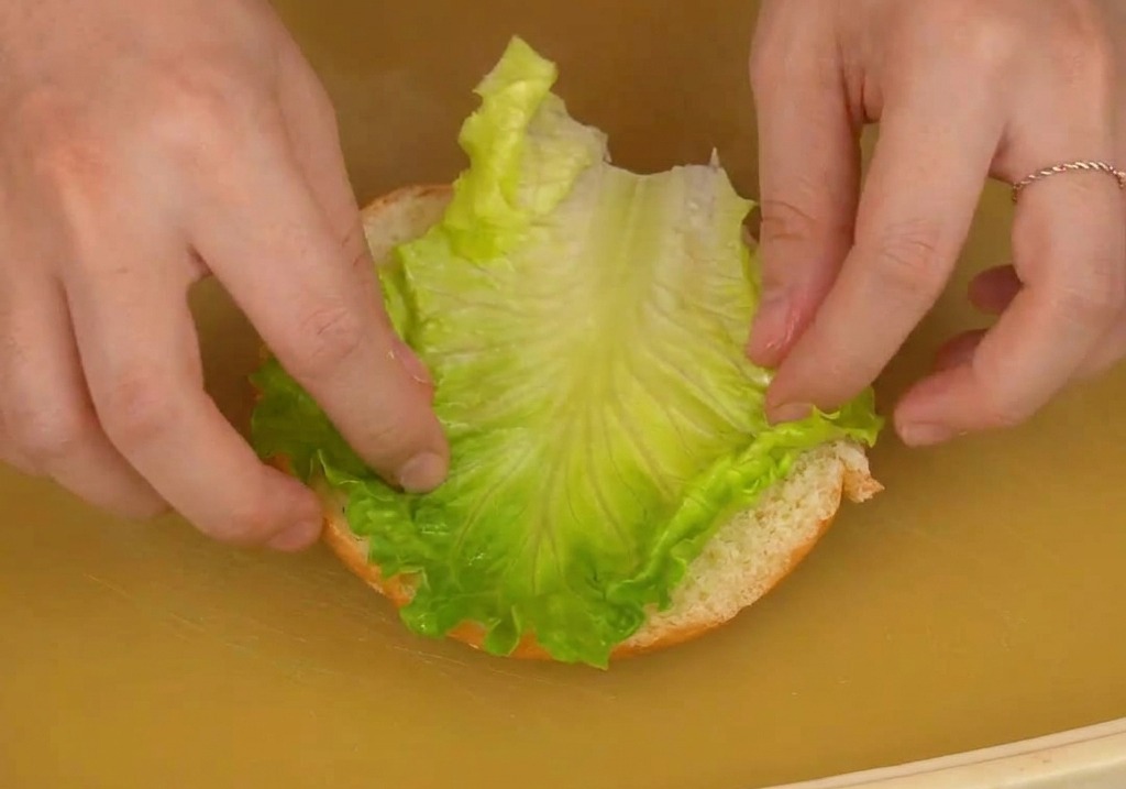 Dwie dłonie kładące świeży liść zielonej sałaty na dolną połówkę bułki do burgera.