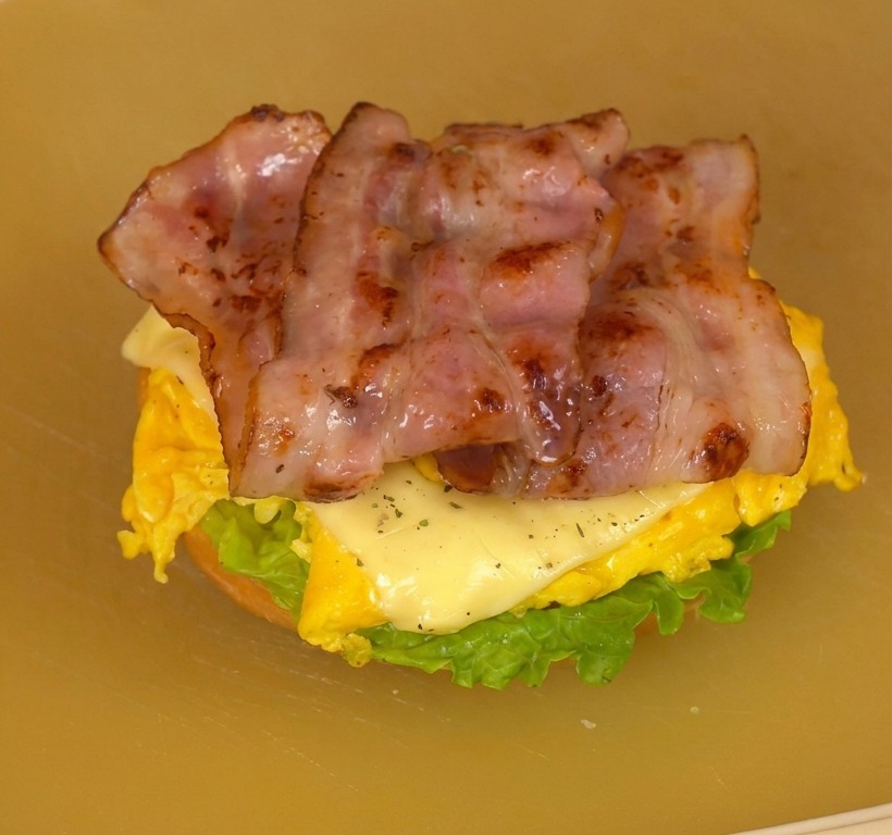 Złożony burger śniadaniowy na desce do krojenia z widocznymi warstwami sałaty, pomidora, jajek i boczku.
