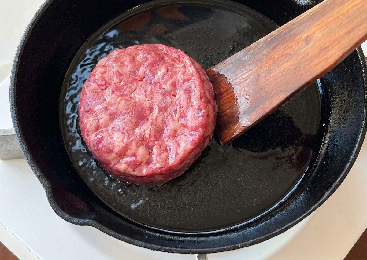 Surowy kotlet wołowy kładziony na żeliwnej patelni za pomocą drewnianej łopatki.