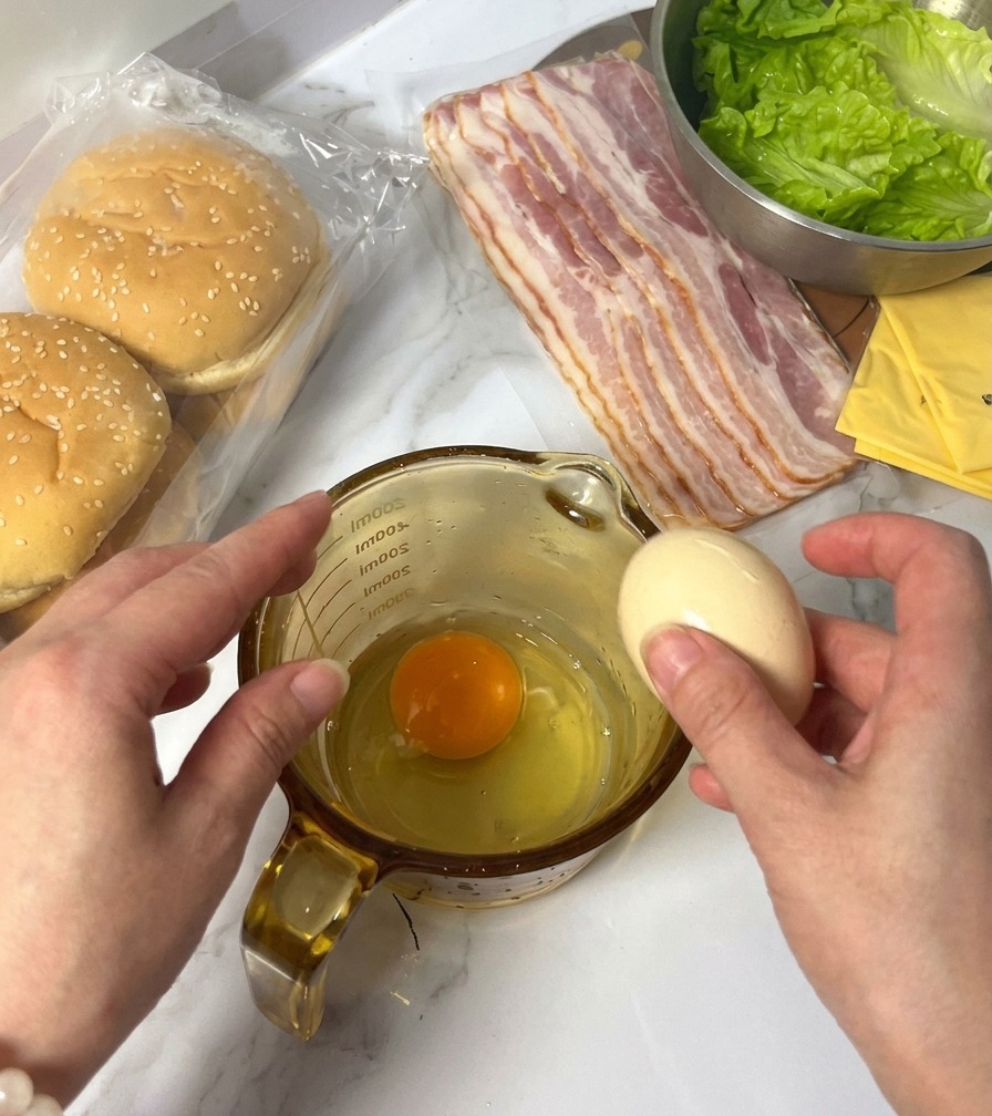 Osoba wbijająca surowe jajko do naczynia z dziubkiem, obok widoczne składniki na burgery.