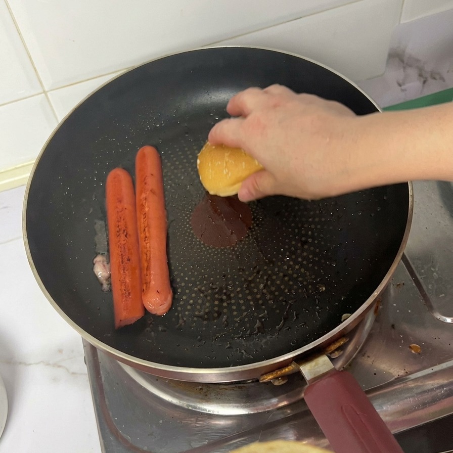 Górna część bułki z sezamem kładziona na patelnię obok rumieniących się parówek.