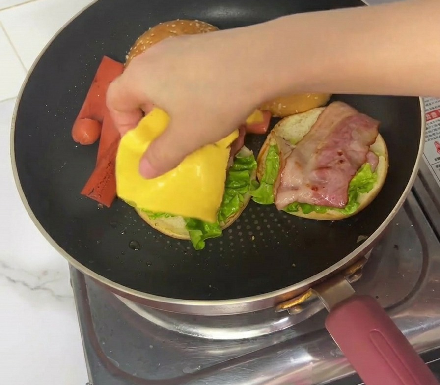 Ręka kładąca plaster sera topionego na składanego burgera na patelni.