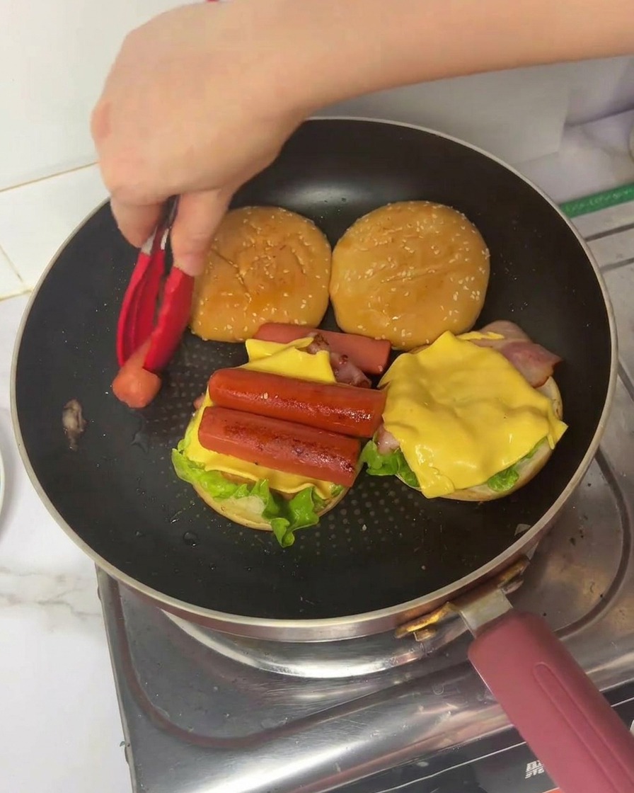 Ręka kładąca złożony kawałek omletu na burgera z boczkiem, serem i parówkami.