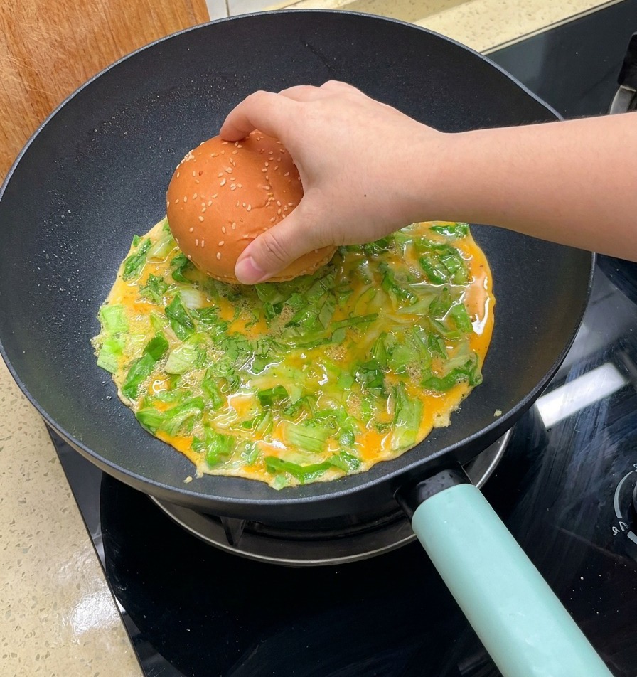 Ręka kładąca połówki bułki z sezamem na smażący się omlet z sałatą na patelni.