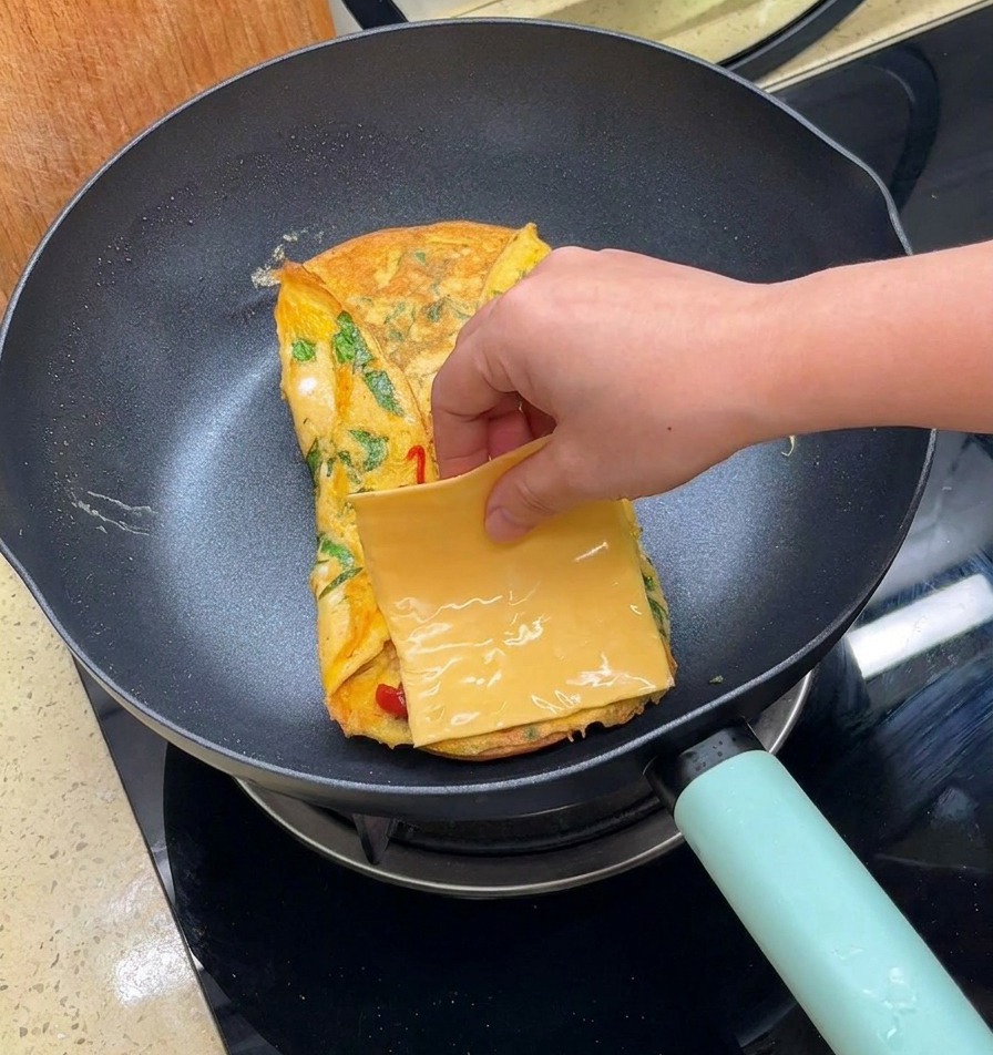 Ręka kładąca plaster żółtego sera na omlet na patelni.