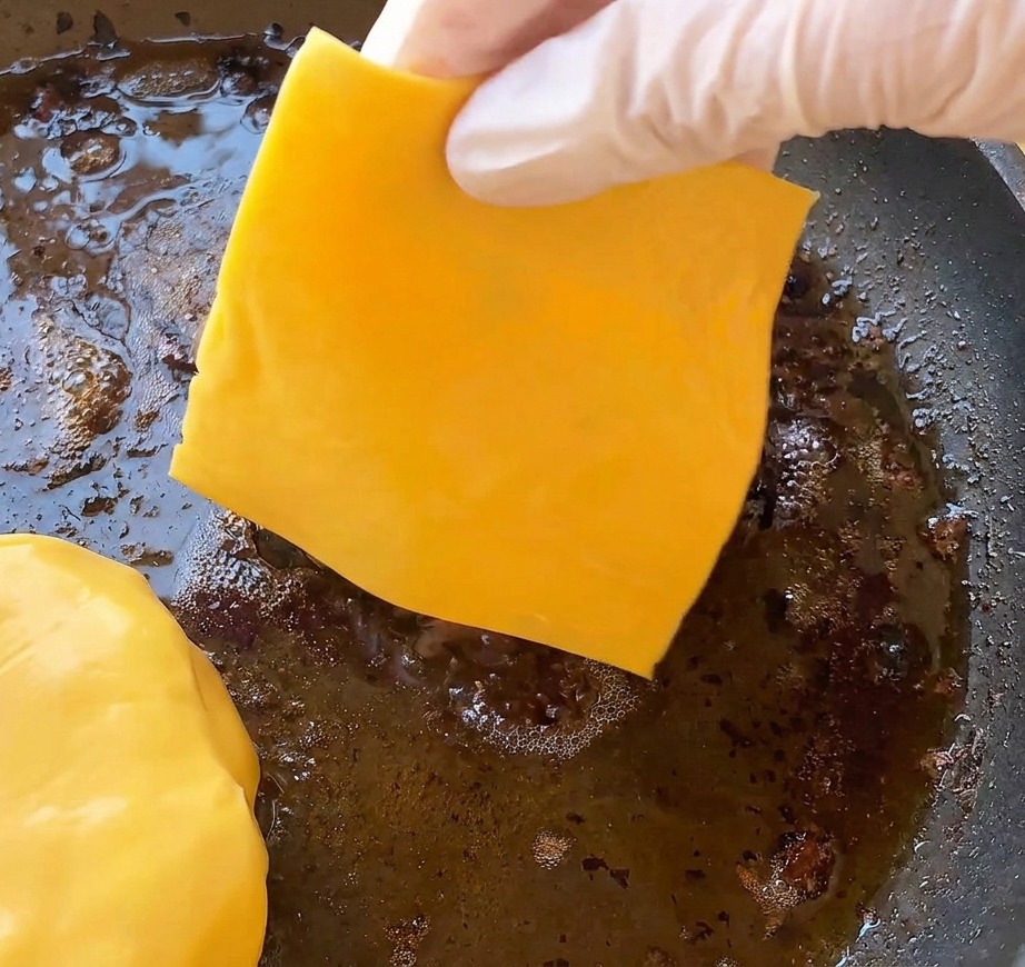 Dłoń w rękawiczce kładąca jaskrawożółty plaster sera cheddar na usmażonym kotlecie na patelni.