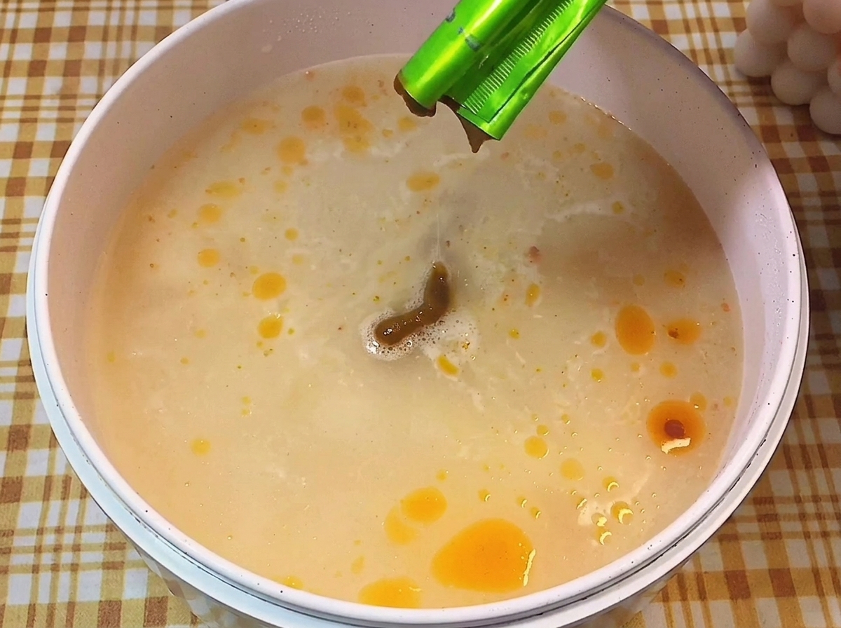 Brązowa pasta przyprawowa wyciskana z zielonej saszetki do garnka z gotującym się, mlecznym wywarem.