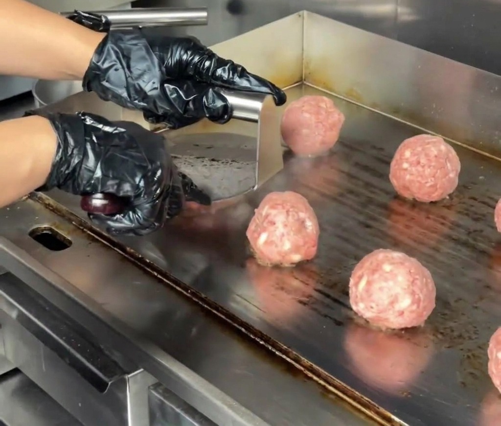 Zbliżenie na stalową prasę do burgerów spłaszczającą kulkę mięsa na gruby kotlet na komercyjnej płycie grzewczej.