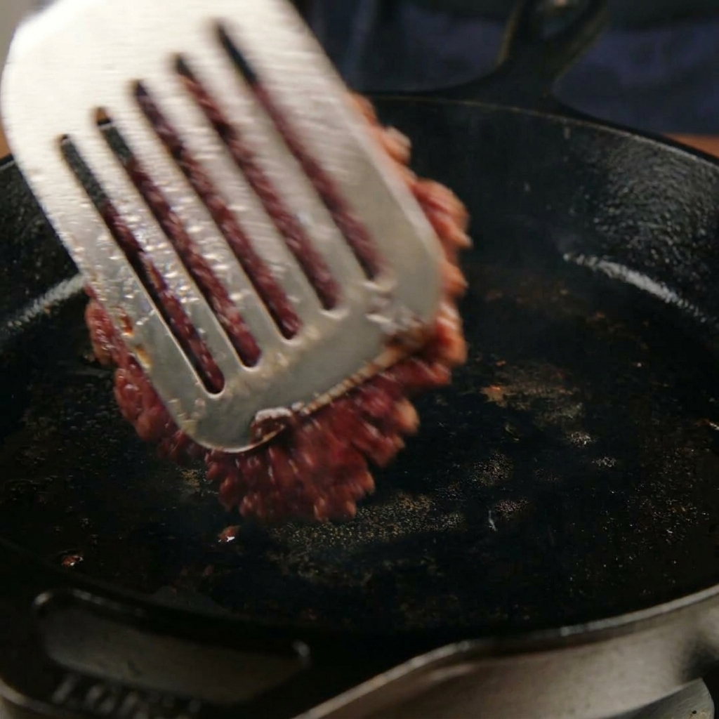 Obracanie mocno skarmelizowanego burgera na patelni za pomocą metalowej łopatki.