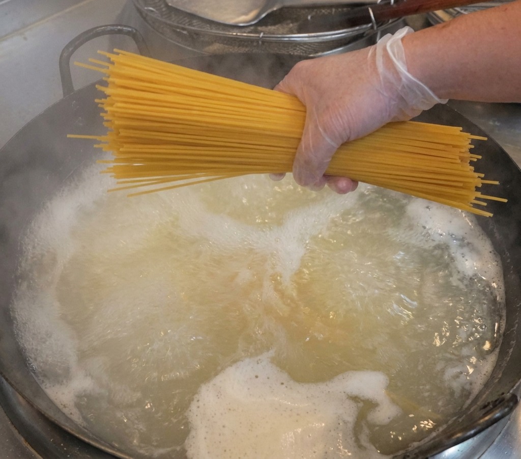 Dłoń w przezroczystej rękawiczce trzymająca wiązkę suchego spaghetti nad garnkiem z wrzącą wodą.