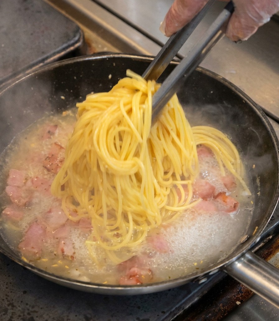 Pęczek żółtego spaghetti podnoszony szczypcami i przekładany na patelnię z kawałkami boczku.