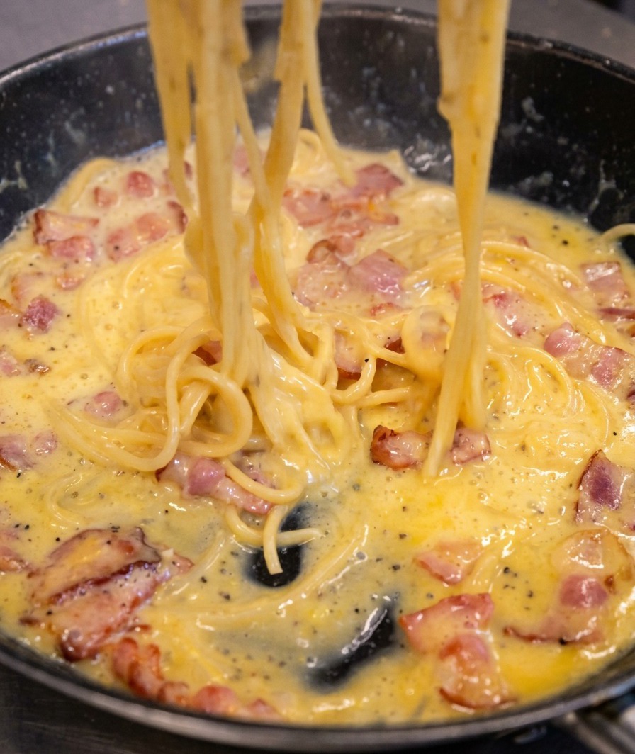 Spaghetti mieszane na patelni, sos zmienia się w gęstą, kremową, bladożółtą powłokę otaczającą makaron.