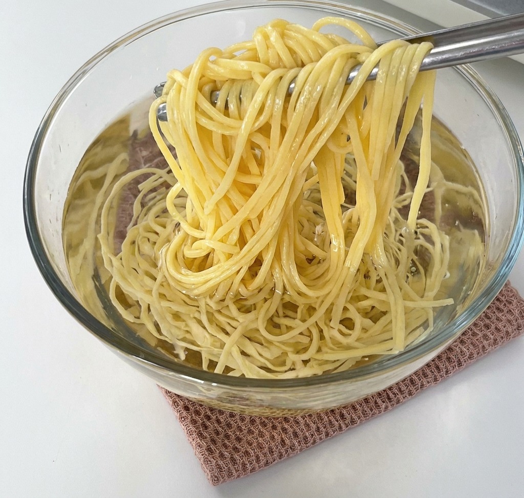 Szczypce podnoszące porcję ugotowanego spaghetti z szklanej miski wypełnionej zimną wodą.