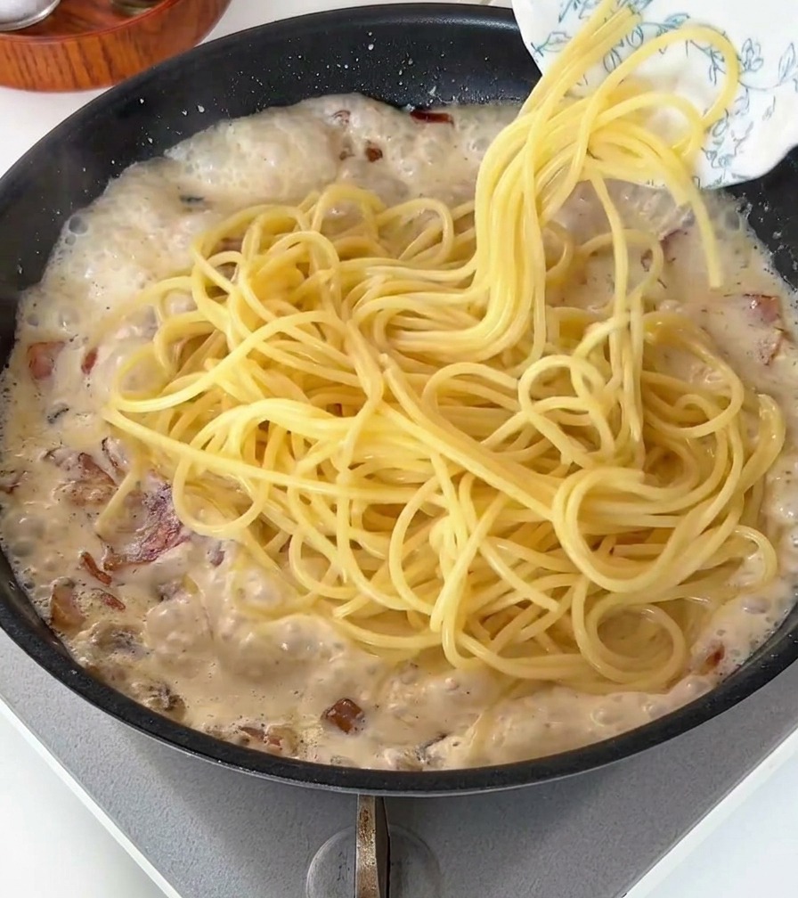 Ugotowany makaron spaghetti wrzucany na patelnię z bąbelkującym, kremowym sosem z boczkiem.