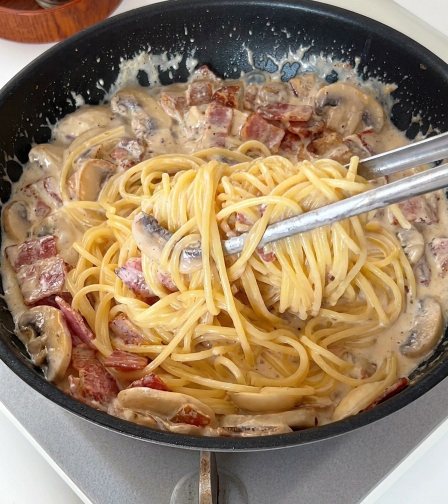Szczypce mieszające makaron spaghetti w gęstym, kremowym sosie z kawałkami boczku i pieczarek.
