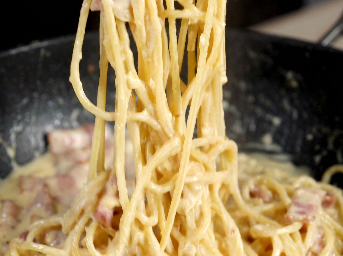 Metalowy młynek do pieprzu mielący ziarna nad patelnią z kremową carbonarą.