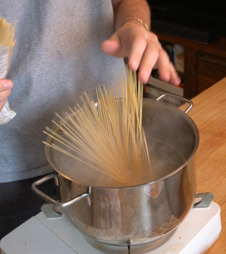 Osoba wrzucająca pęczek spaghetti do garnka z wrzącą wodą na przenośnej kuchence.