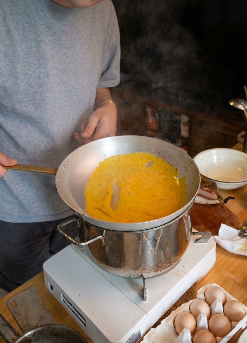 Szef kuchni mieszający spaghetti na metalowej patelni trzymanej nad parującym garnkiem z wodą.