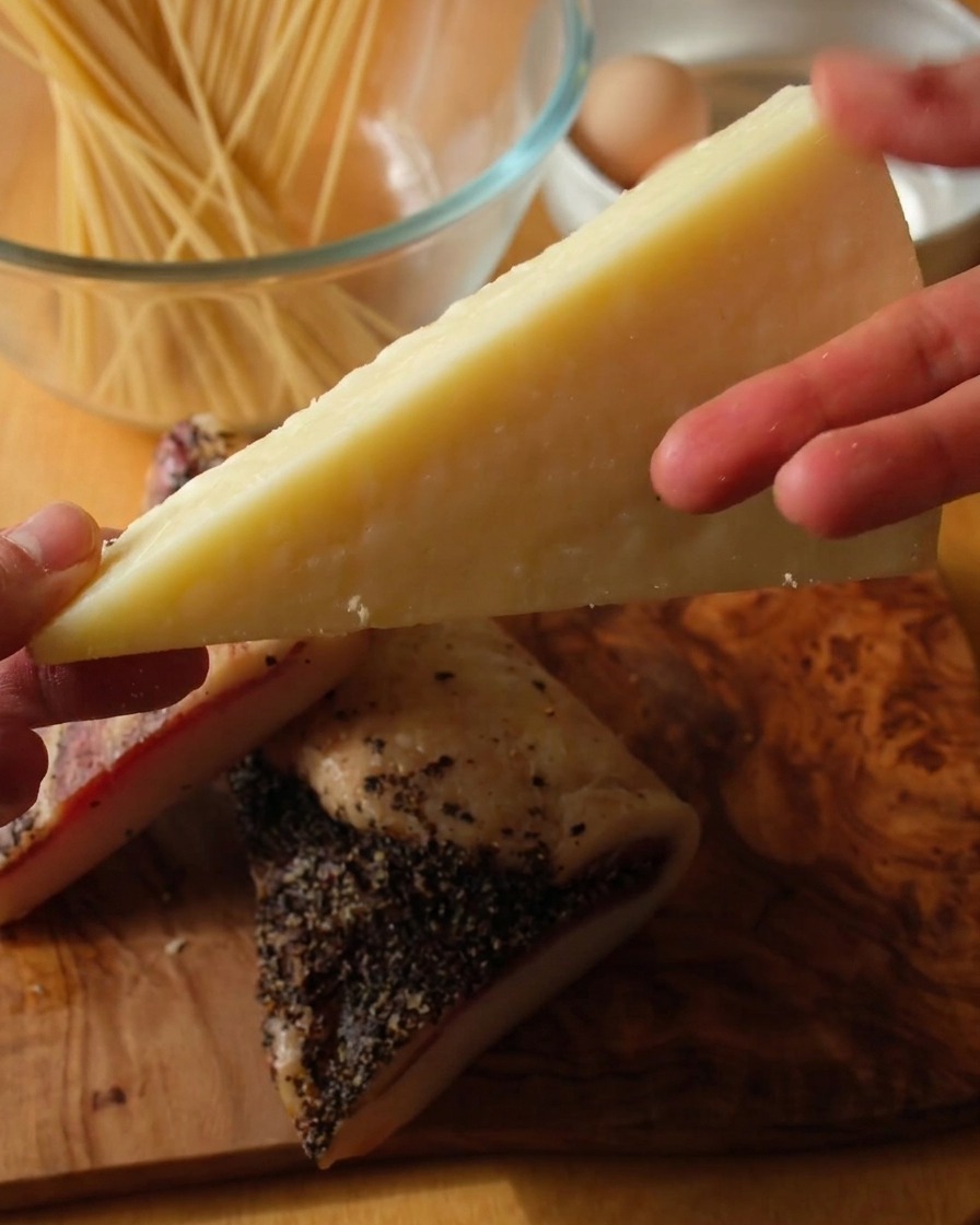 Zbliżenie na dłoń trzymającą kawałek sera Pecorino Romano nad dwoma grubymi plastrami guanciale w posypce z pieprzu na drewnianym blacie.