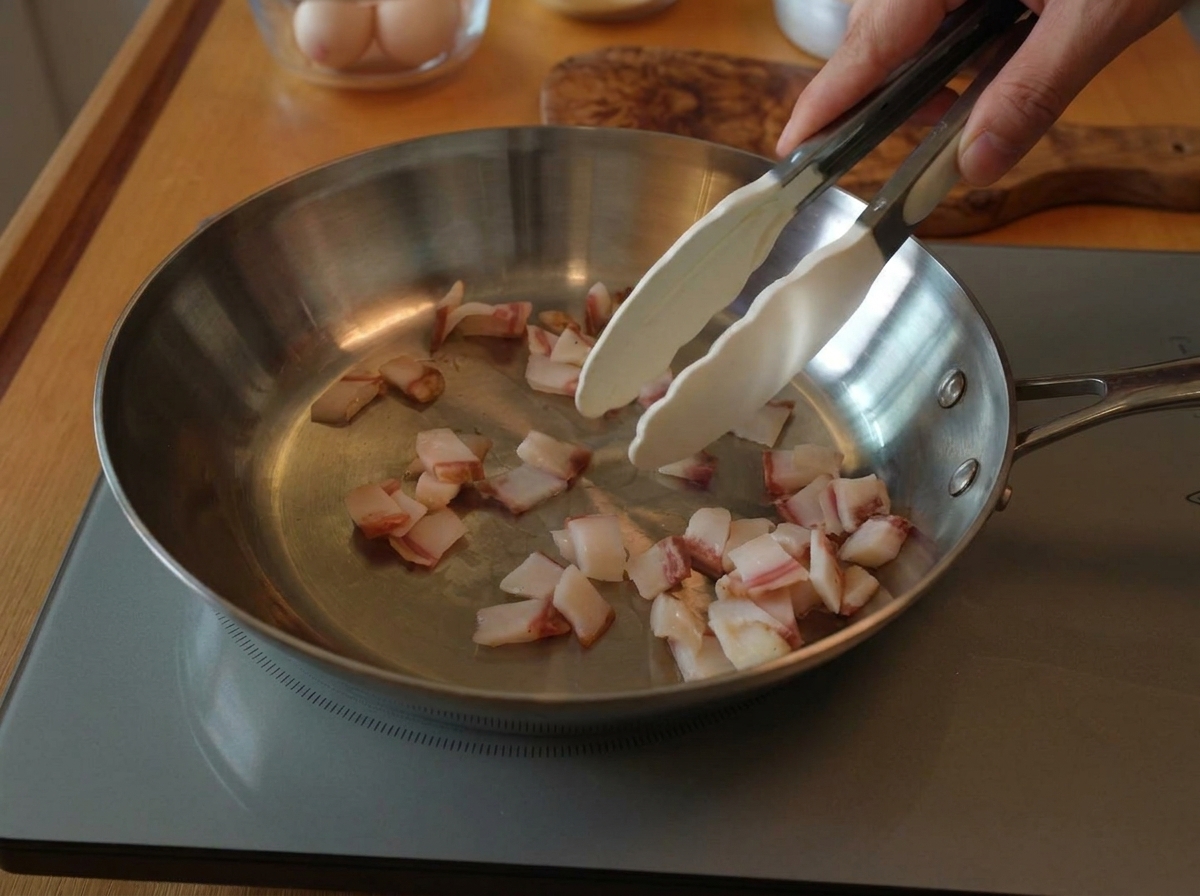Szczypce mieszające kawałki surowego guanciale na szerokiej patelni ze stali nierdzewnej na płycie indukcyjnej.