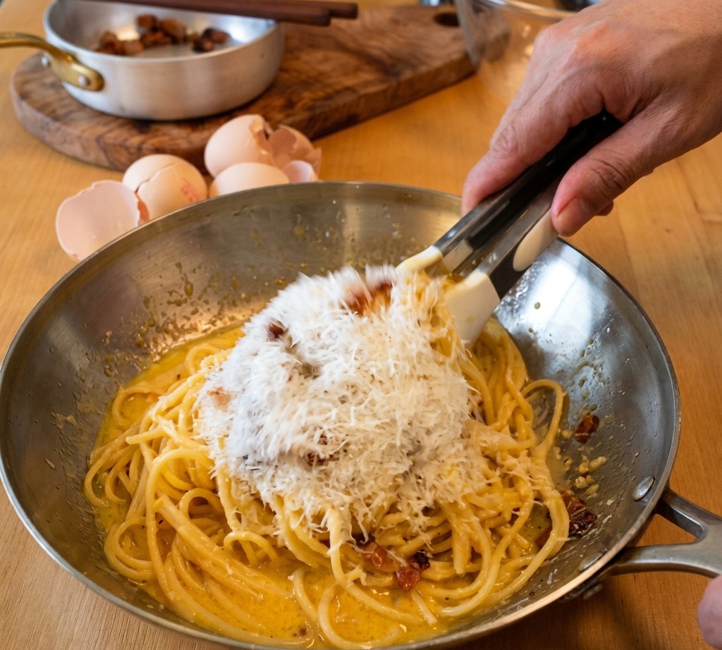 Szczypce mieszające spaghetti na patelni, podczas gdy do kremowego sosu jajecznego dodawana jest duża ilość drobno startego sera Pecorino Romano.