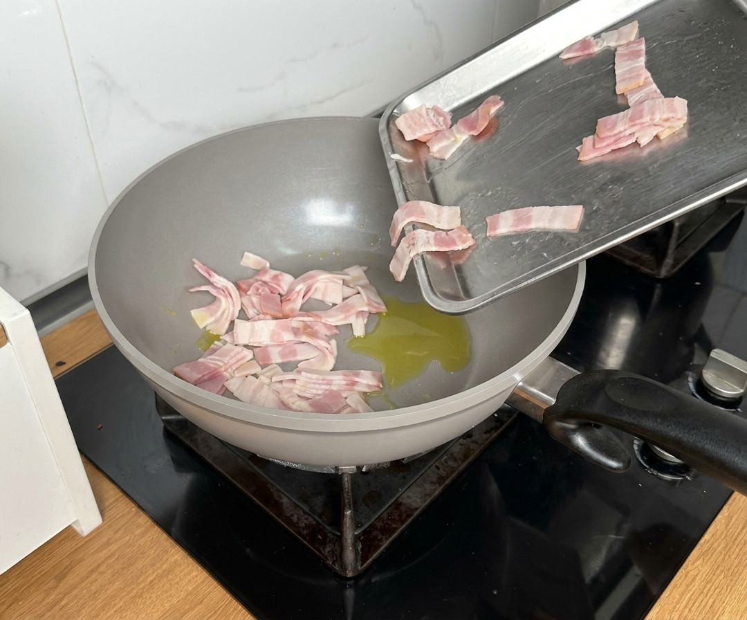 Kawałki boczku wrzucane z metalowej tacy na dużą patelnię z rozgrzaną oliwą.