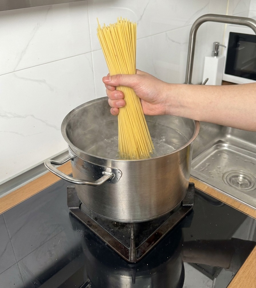 Pęczek suchego spaghetti wkładany do stalowego garnka z wrzącą wodą.