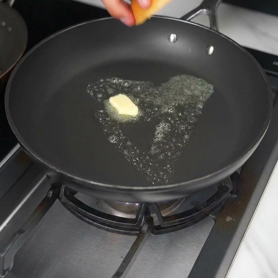 Ręka kładąca bułkę brioche na patelnię z roztopioną kostką masła.