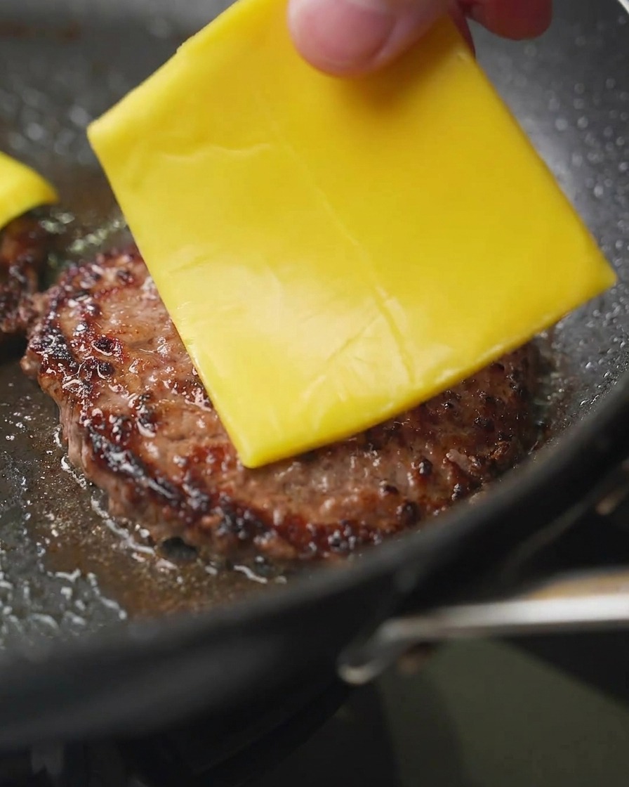 Zbliżenie na kładzenie plastra żółtego sera cheddar na podsmażony, brązowy kotlet wołowy.