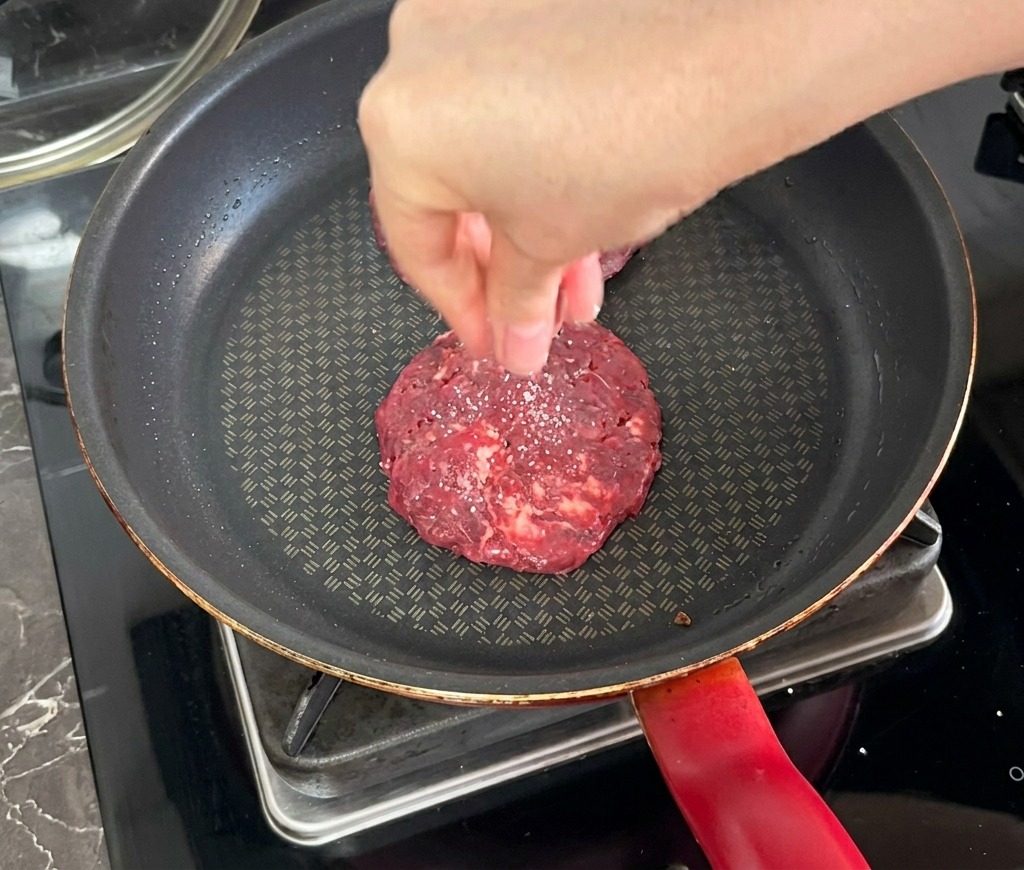 Dłoń posypująca solą smażony kotlet wołowy na ciemnej patelni.