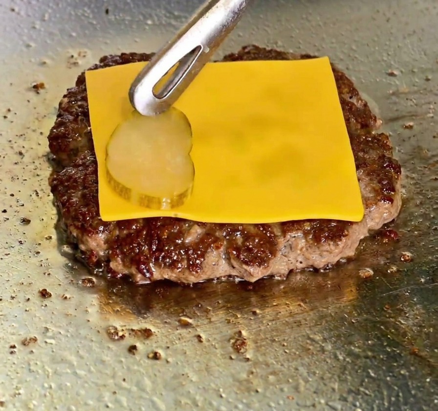Roztopiony ser cheddar na mięsie i plasterek ogórka korniszona kładziony szczypcami.