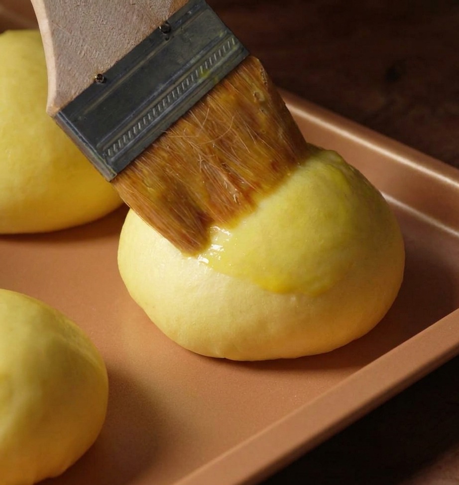Pędzel cukierniczy nakładający lśniącą glazurę z jajka na puszystą bułkę brioche.