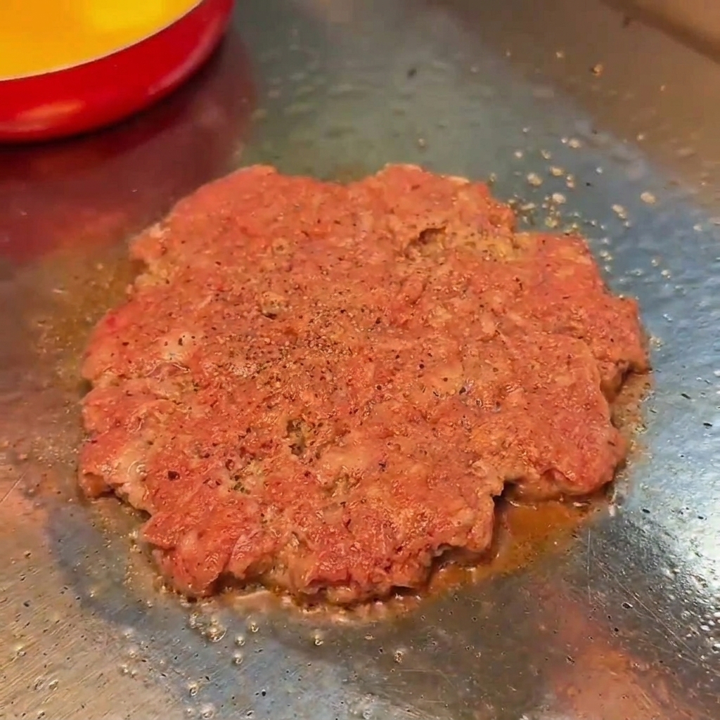 Kotlet wołowy smażący się na płaskim grillu, posypany solą i czarnym pieprzem.