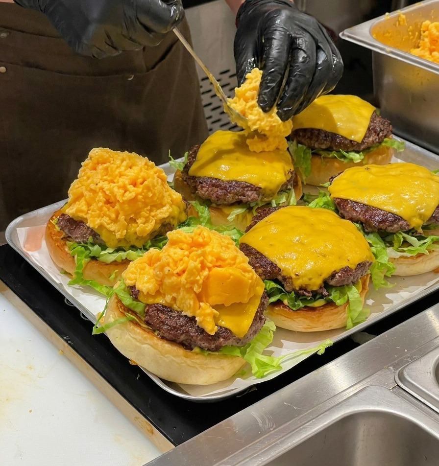 Szef kuchni nakładający łyżką dużą porcję aksamitnej jajecznicy na cheeseburgery.