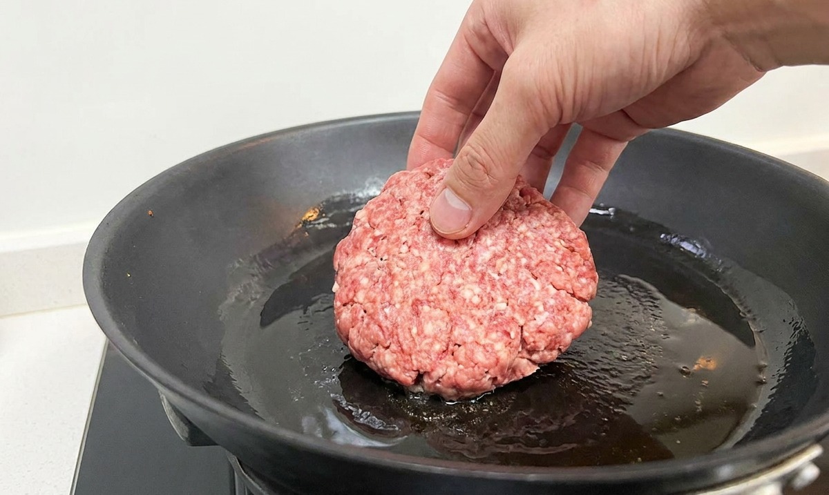 Dłoń kładąca okrągły kotlet z surowej wołowiny na gorącą, czarną patelnię z mieniącym się olejem.