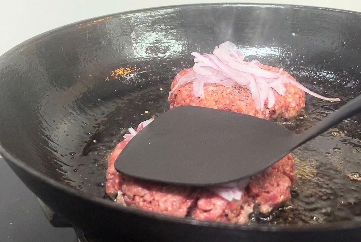 Czarna metalowa łopatka dociskająca kotlet wołowy z cebulą do gorącej patelni.