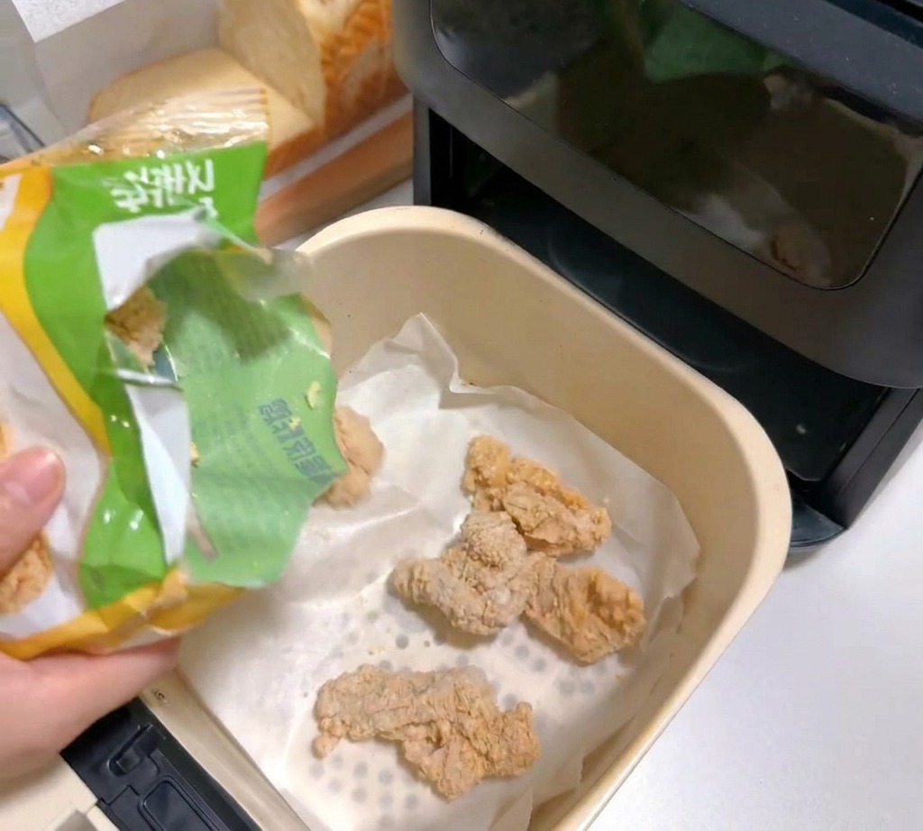 Mrożone kawałki kurczaka w panierce wsypywane do koszyka frytkownicy wyłożonego papierem do pieczenia.