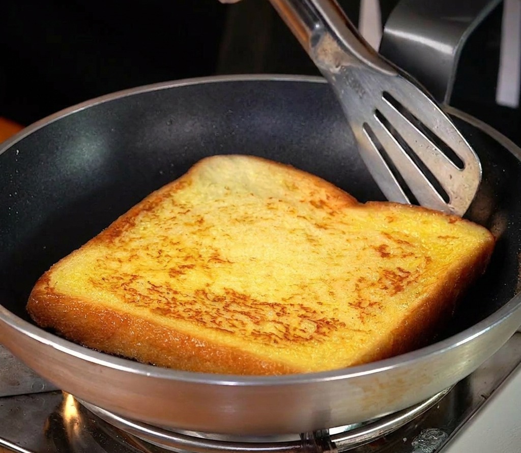 Pojedyncza kromka grubego tosta smażona na srebrnej patelni z masłem, widoczna rumiana skórka.