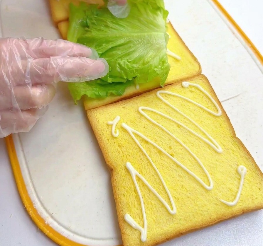 Dłonie kładące świeży liść sałaty na kromkę tosta polaną sosem.