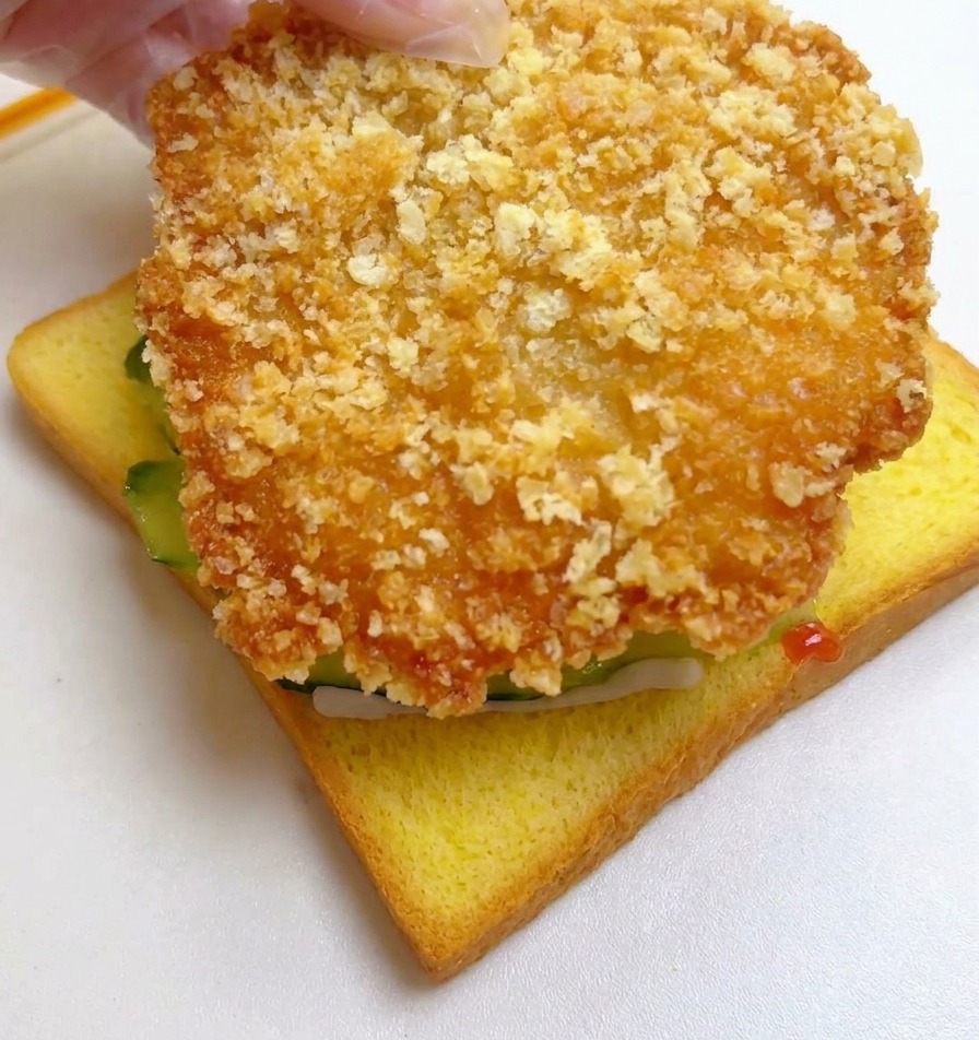 Dłoń kładąca duży, usmażony na złoto filet z kurczaka na kromkę tosta z ogórkiem.