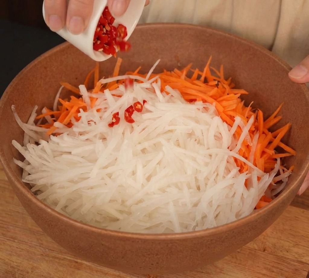 Miska z pokrojoną w słupki białą rzodkwią i marchewką, do której dodawane jest posiekane chili.